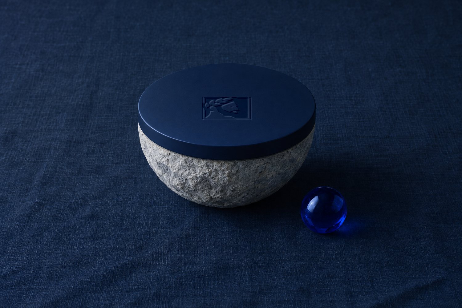A rough stone bowl capped with a smooth navy lid pressed with the Olivent mark, a cobalt glass sphere beside it