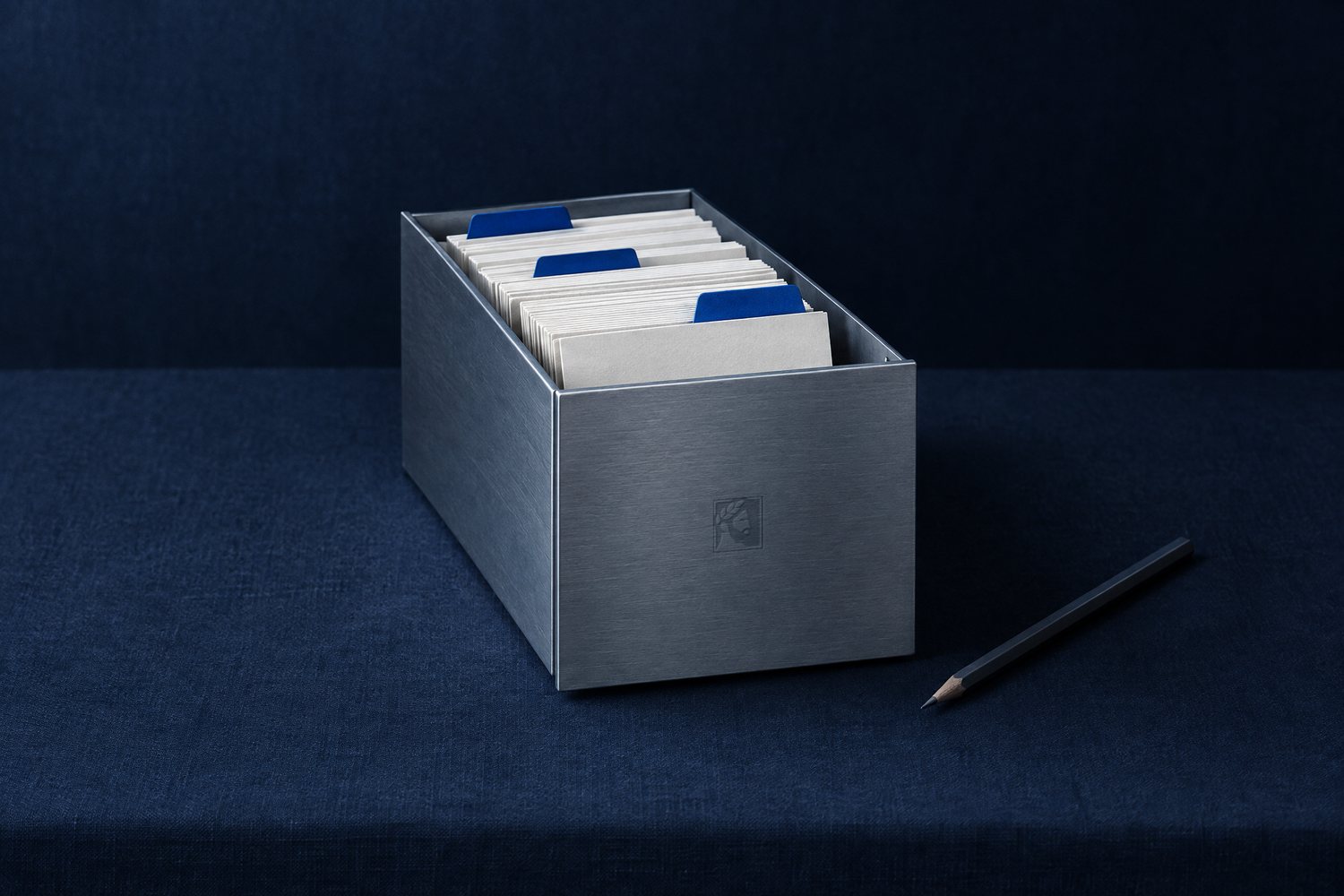 A brushed steel card-file box of blank index cards with cobalt dividers and a pencil, the Olivent mark engraved on its front