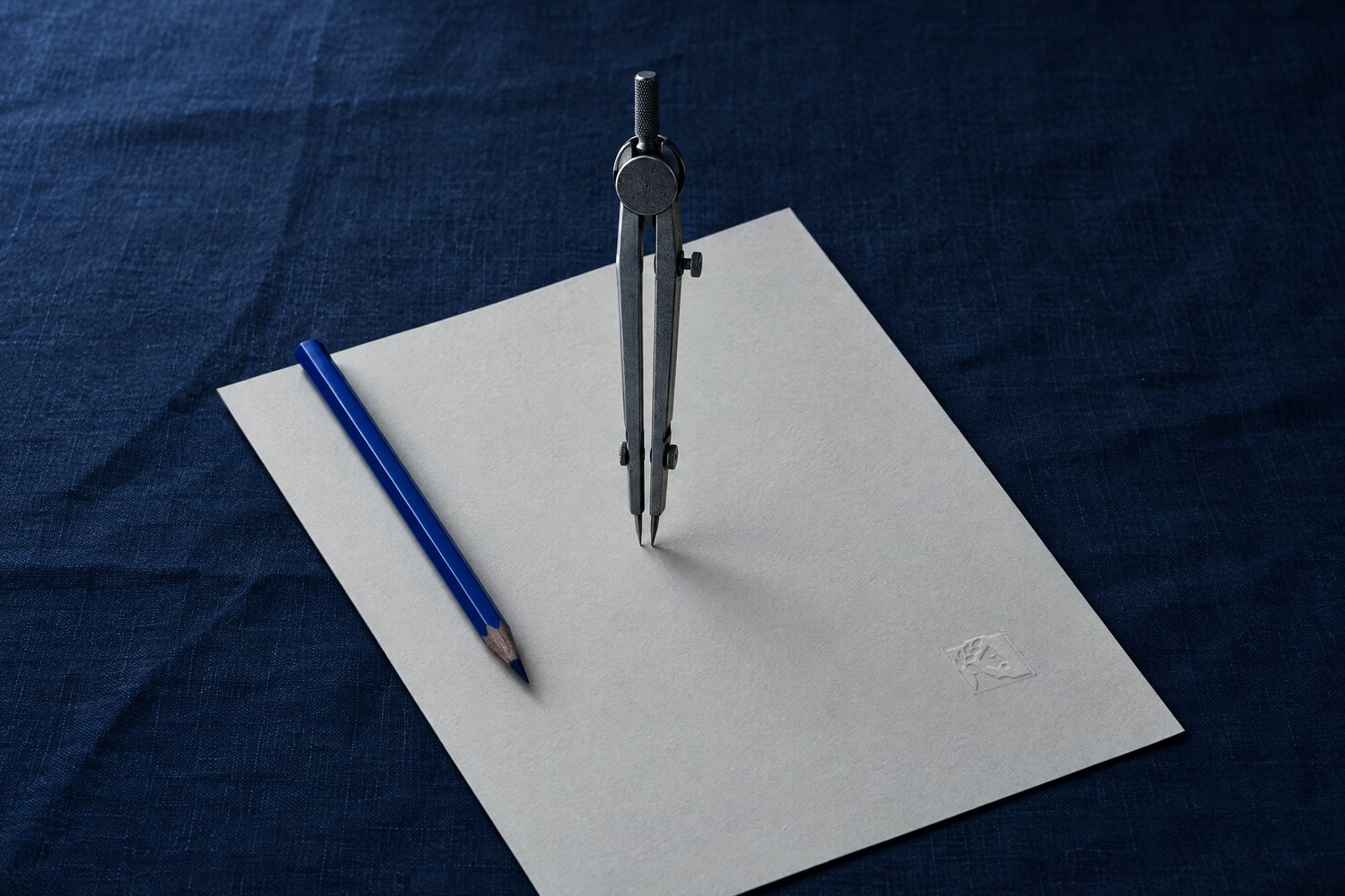 A steel drafting compass with its legs drawn close on a sheet of paper embossed with the Olivent mark, a cobalt pencil beside it