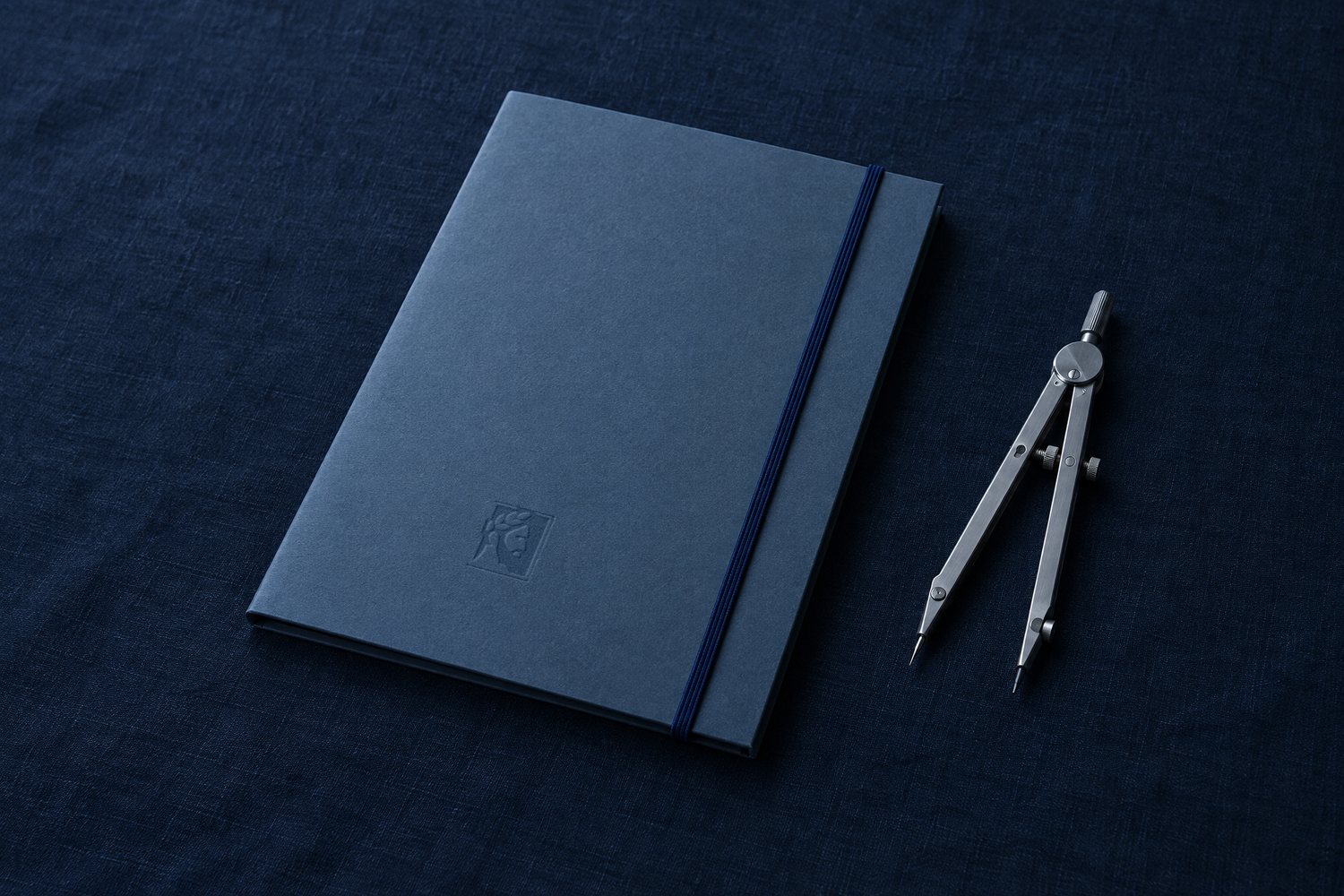 A closed slate blue folder with a navy elastic cord beside a steel drafting compass, the Olivent mark pressed into the cover
