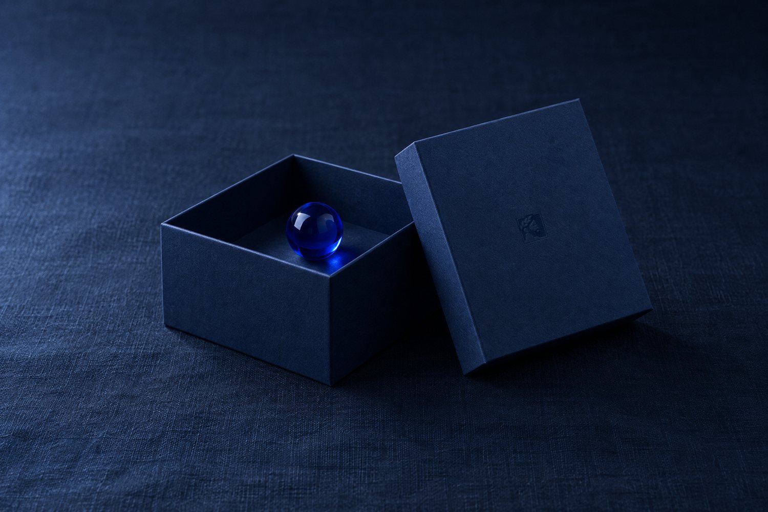 An open navy box holding a cobalt glass marble, its lid leaning beside it with the Olivent mark pressed into the lid